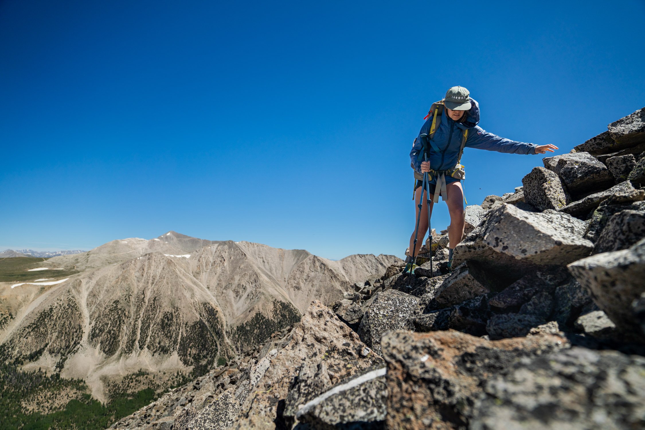 A person climbing rocky terrain on a mountain trail under a clear blue sky, wearing outdoor gear and carrying a backpack.