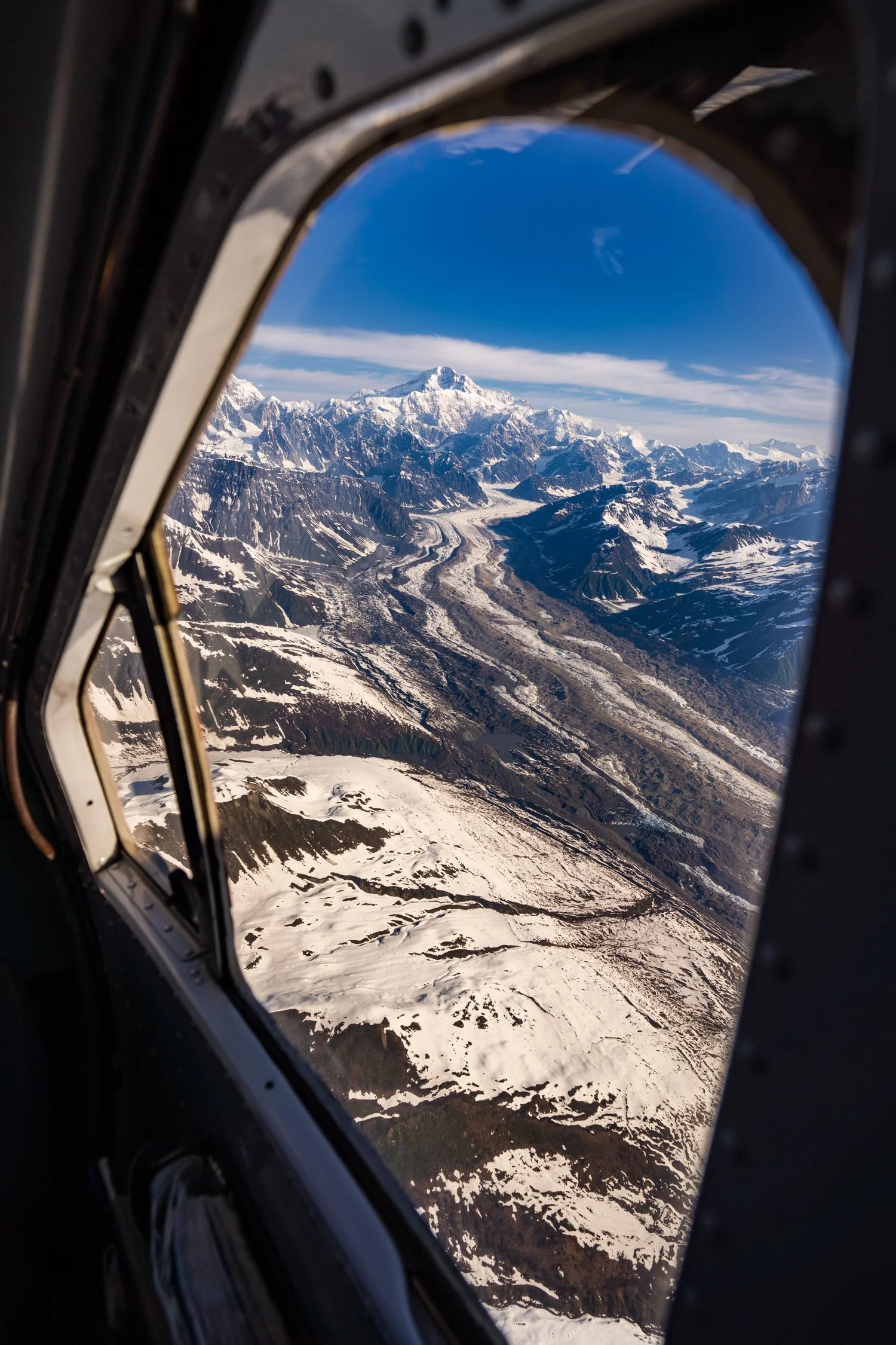 Snow-covered mountain range and glaciers seen through an aircraft window, with the tallest peak in the center topped with snow and surrounded by rugged, icy terrain.