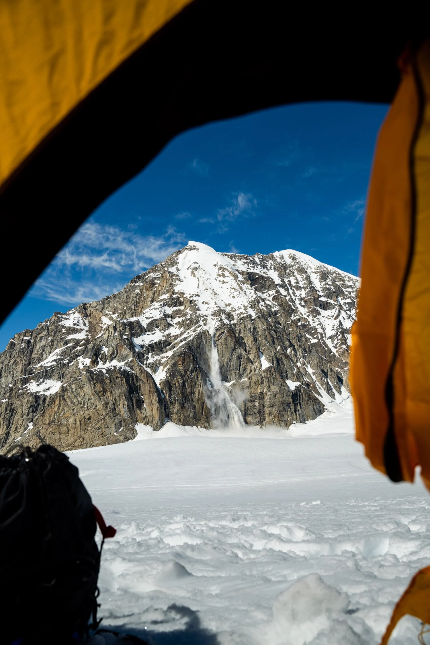 Photo of a snow-covered mountain with a waterfall cascading down, taken from inside a yellow tent. The sky is blue with some clouds.