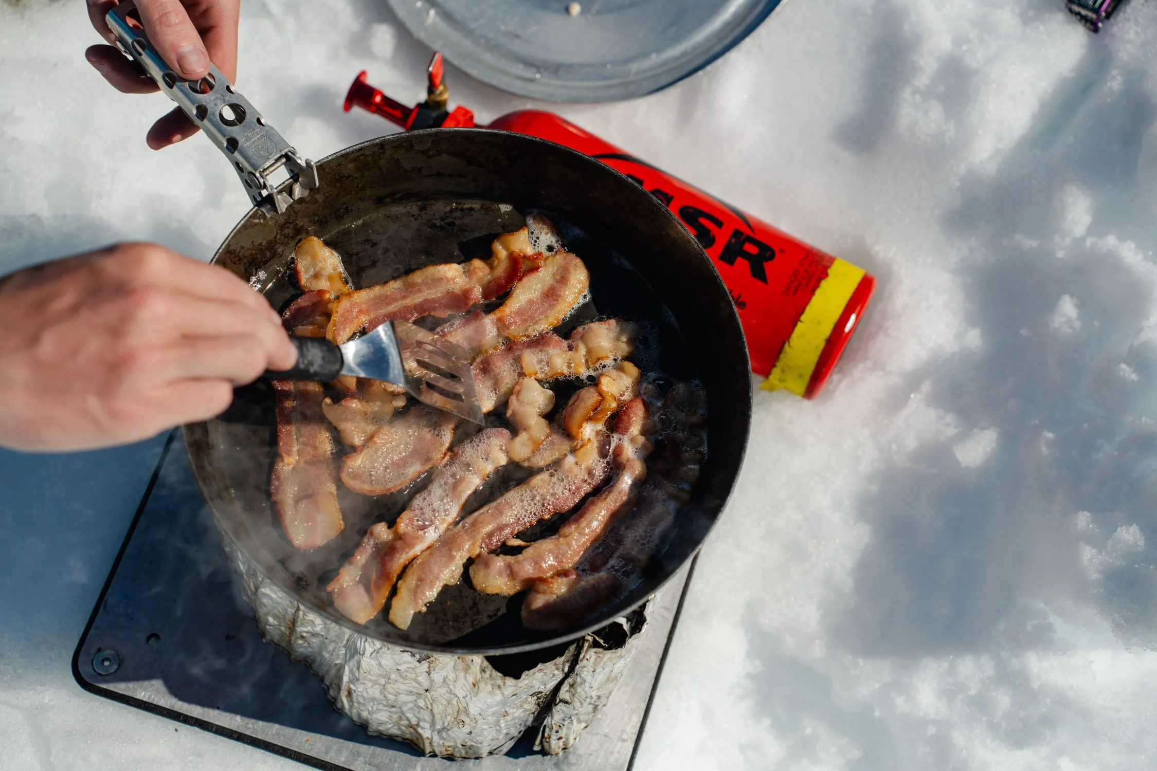 Bacon cooking in a frying pan over a portable propane stove on snow.