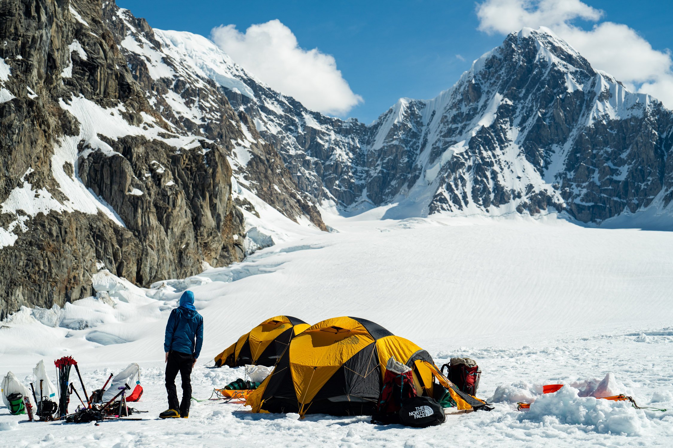 A person in a blue jacket and black pants standing near yellow tents in a snowy mountain landscape with rocky cliffs and snow-capped peaks.