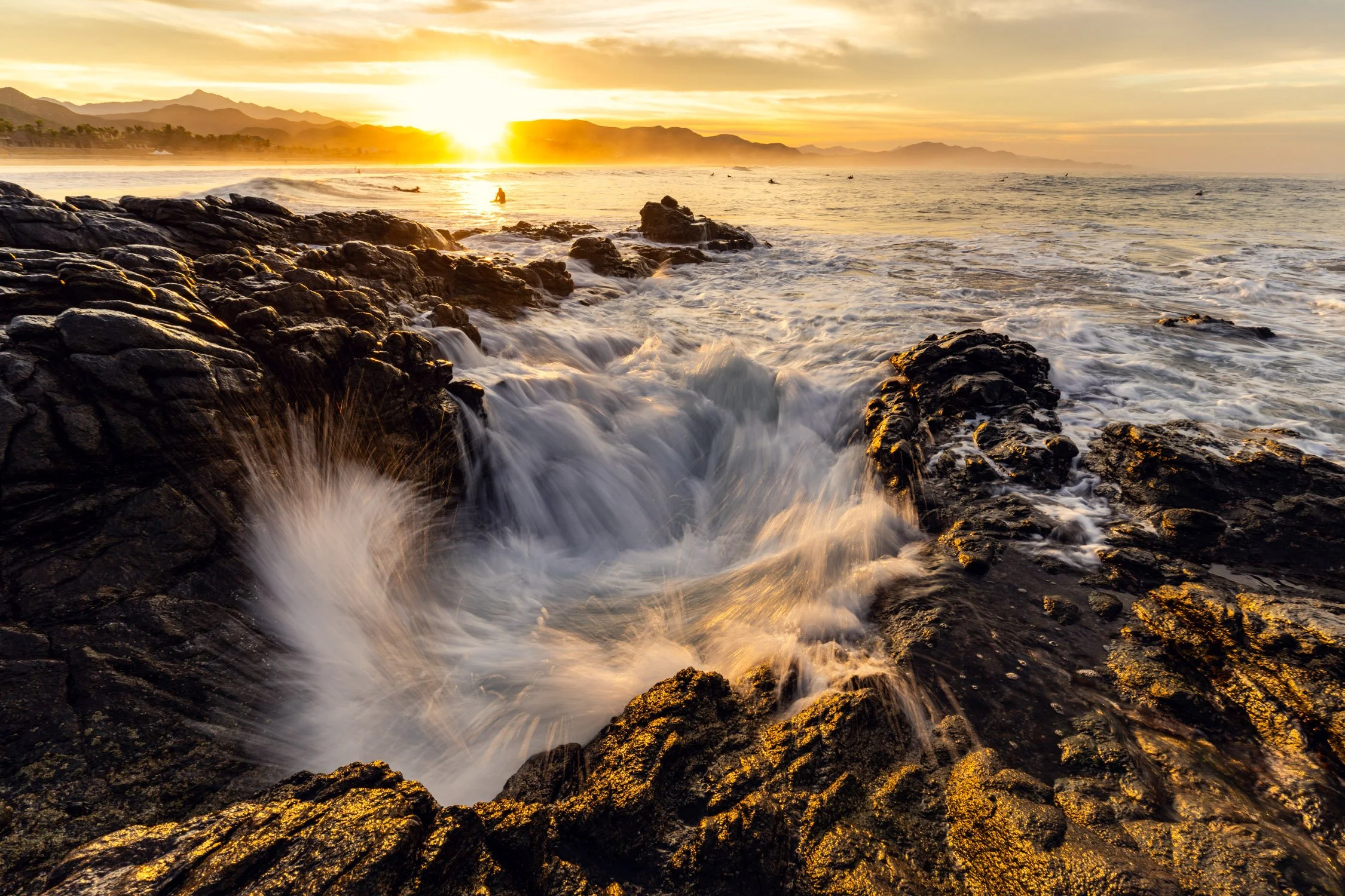 Outdoor photographer Dalton Johnson captures an image of a wave splashing on the rocks at Cerritos Beach with surfers in the background