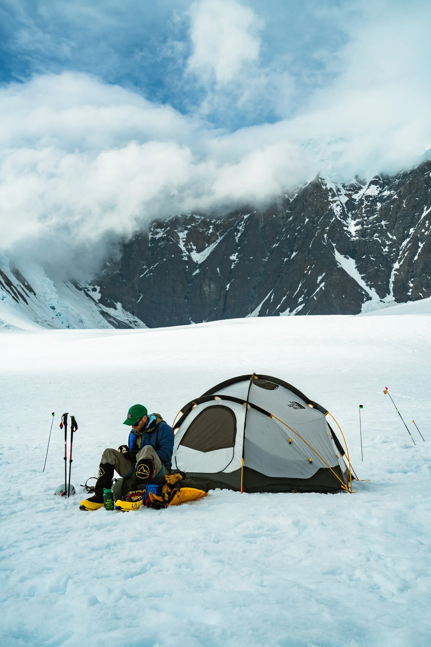 A person sitting beside a camping tent on snow, surrounded by mountaineering gear, with snow-covered mountains and clouds in the background.