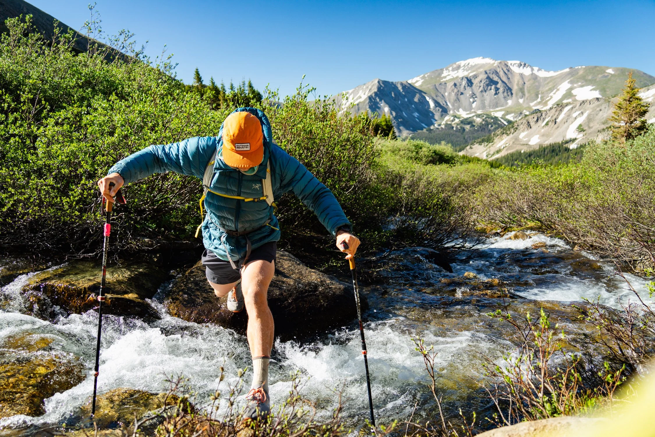 Hiker crossing a stream in a mountainous landscape with snow-capped peaks in the background.