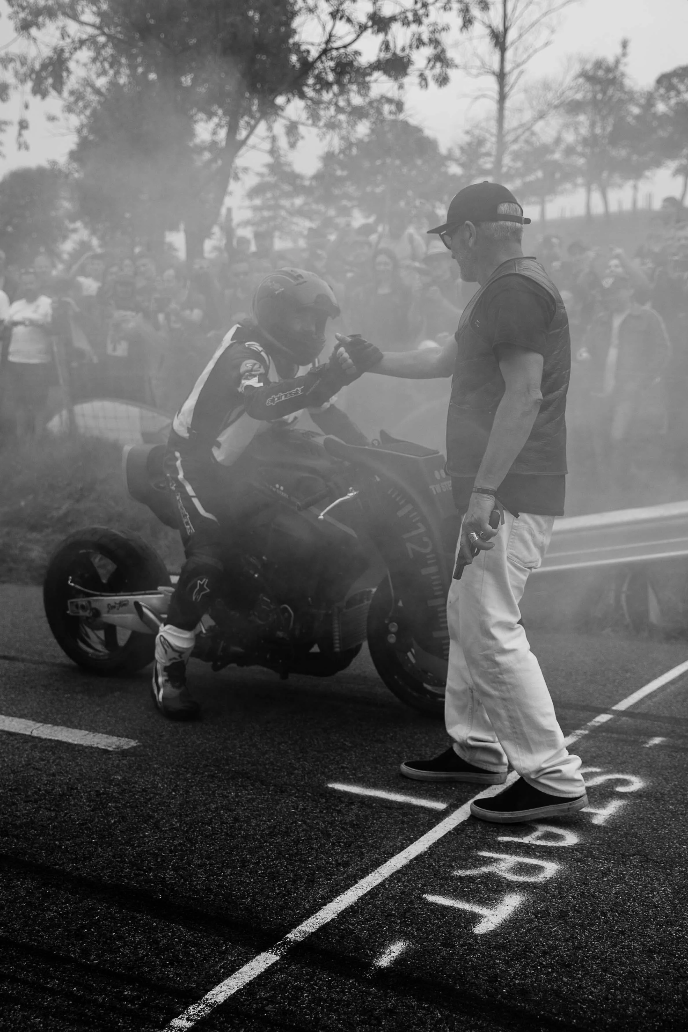 A motorcyclist wearing racing gear and helmet is receiving a handshake from a man in casual clothing at a race track. The scene is smoky with many onlookers in the background.