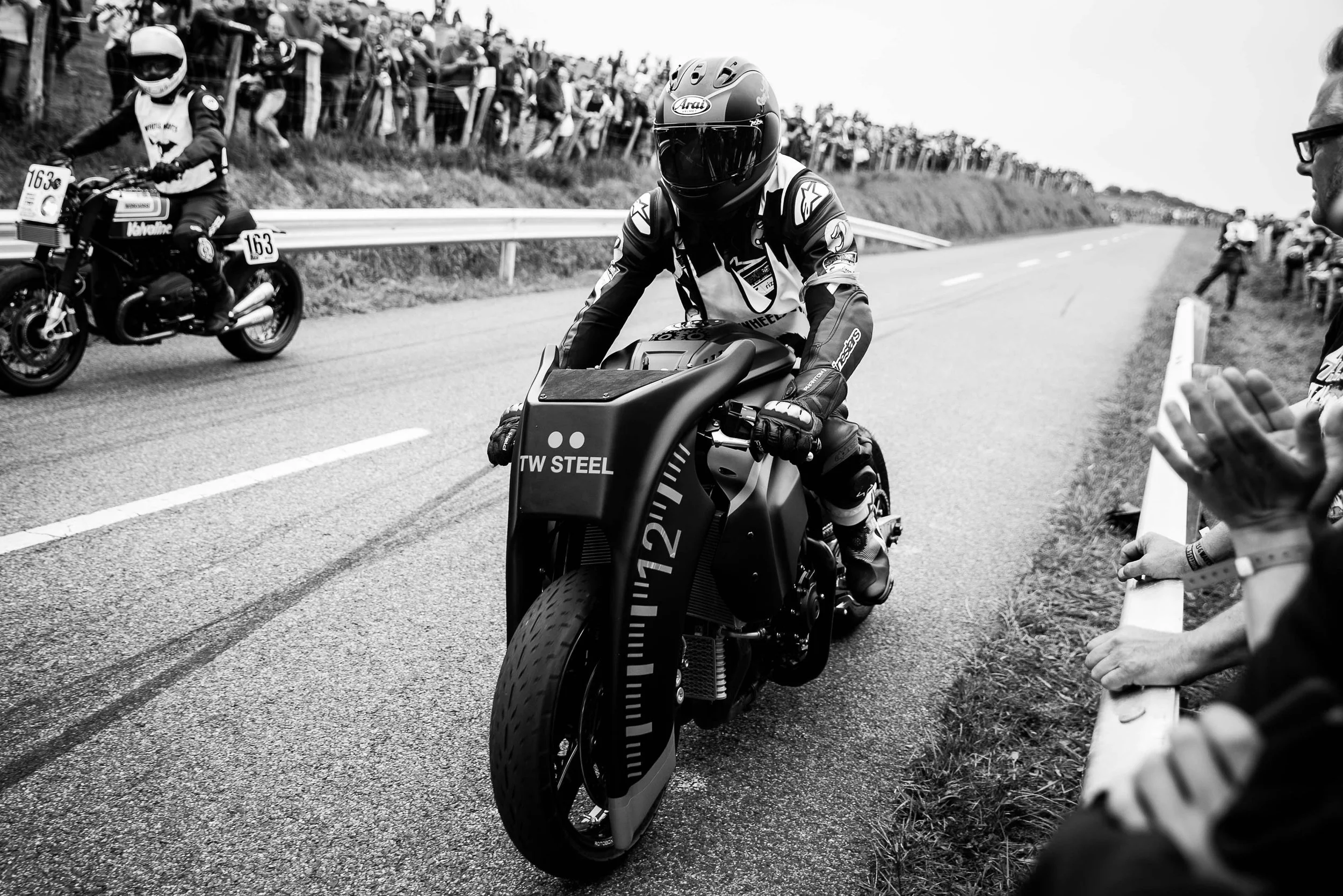A motorcycle racer in a helmet and racing gear sitting on a black racing motorcycle on a road, with spectators behind a guardrail watching on both sides of the road during a race.