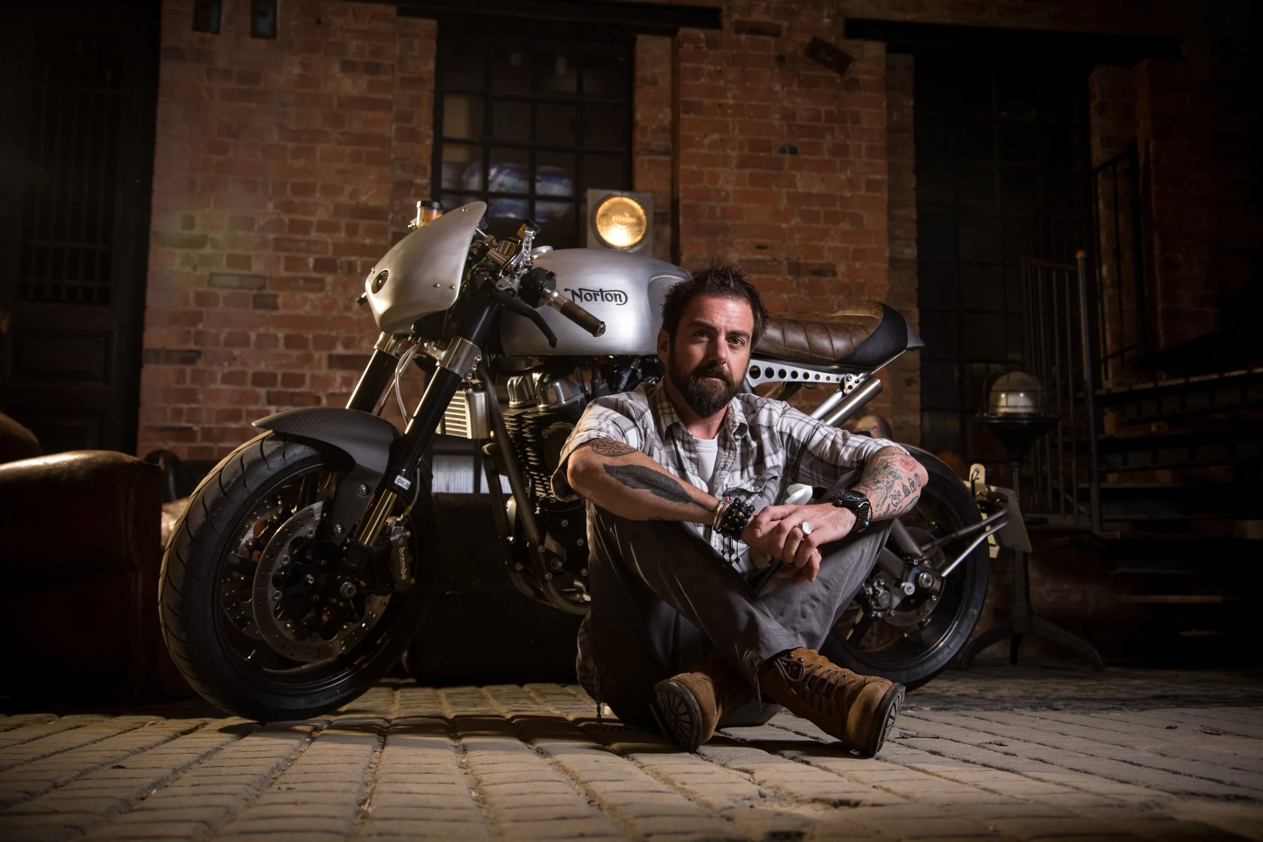 A man sitting cross-legged on a brick floor next to a custom motorcycle inside an industrial-style room with exposed brick walls, a window, and metal staircase.