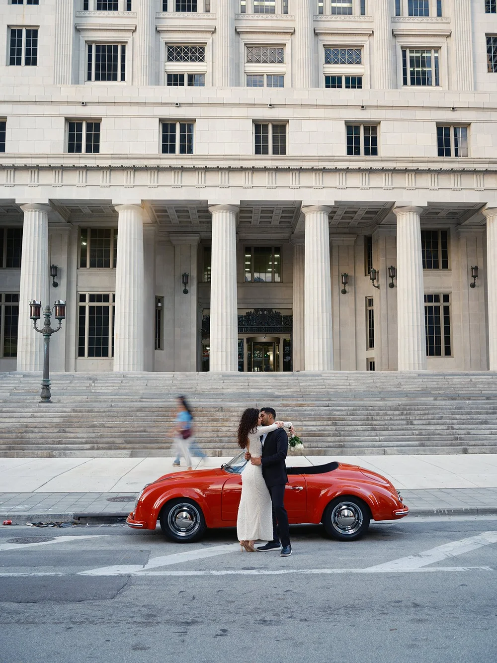 Bride and groom standing by a red vintage convertible in front of the columned Miami-Dade County Courthouse in downtown Miami