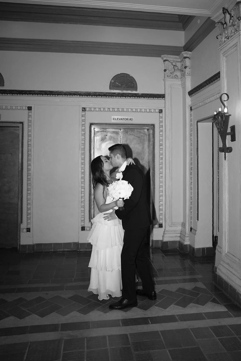 Black and white wide shot of the couple kissing in a hallway with a patterned tile floor