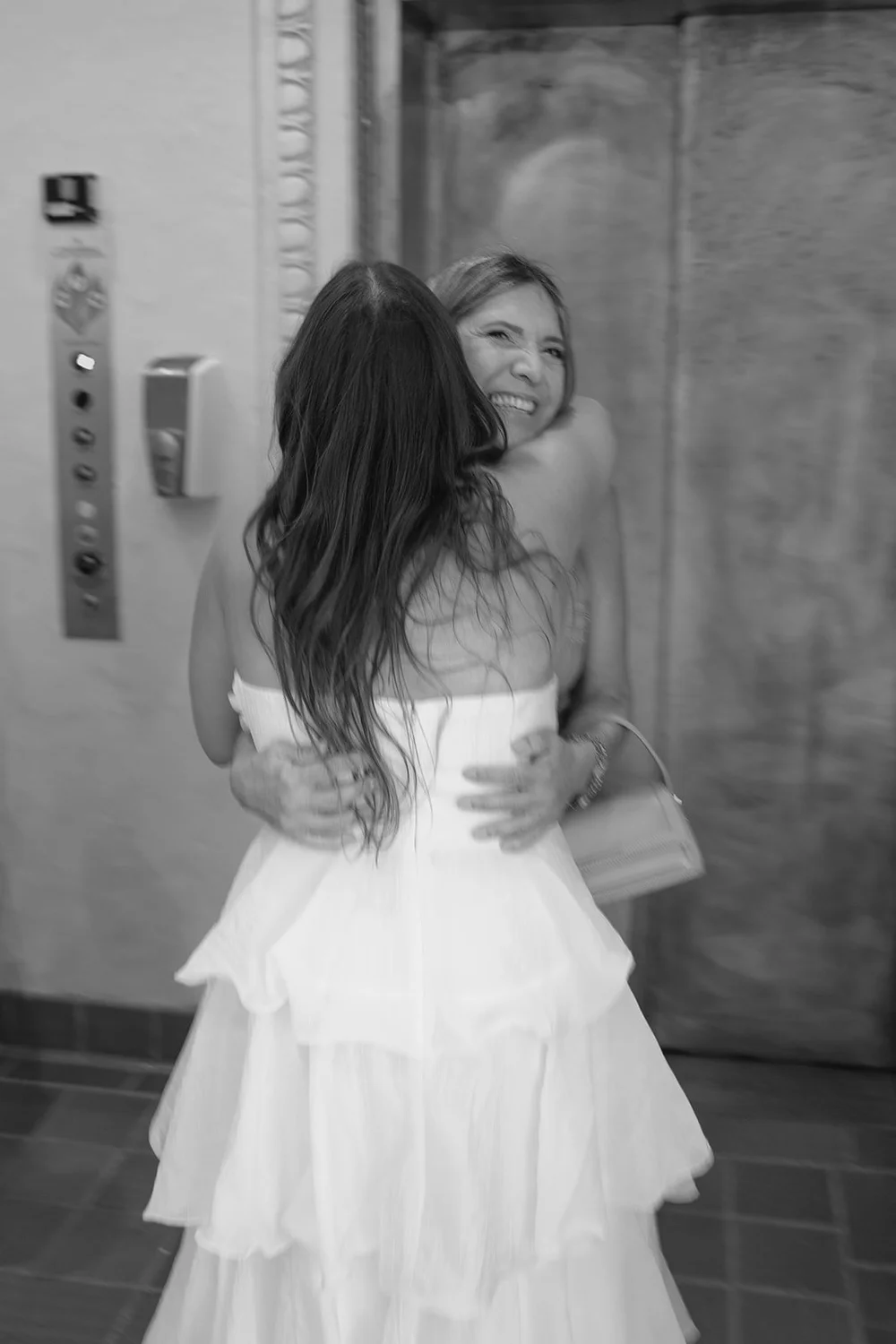 Black and white image of Chris from behind, her tiered ruffle dress, hugging a guest in the hallway