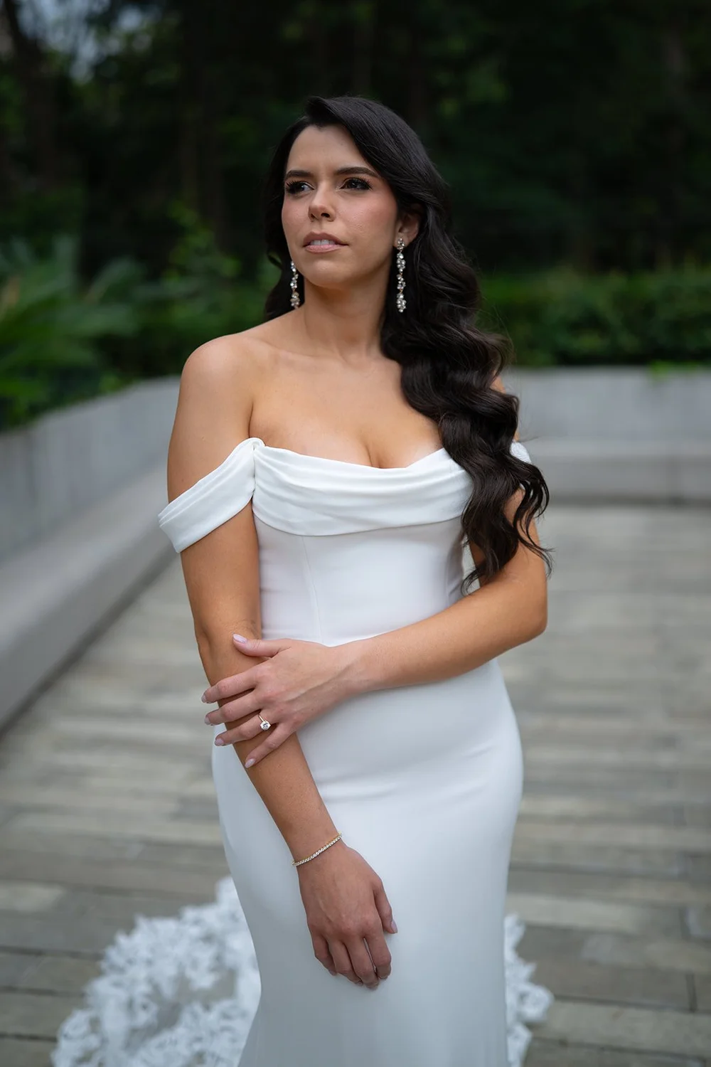 Krystine in a white off-the-shoulder gown standing on a wooden terrace with greenery behind her