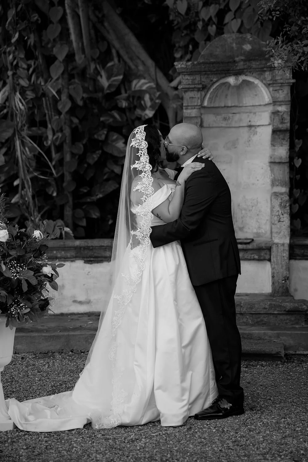 Maria and Christian kissing in front of the stone niche with greenery around them, in black and white