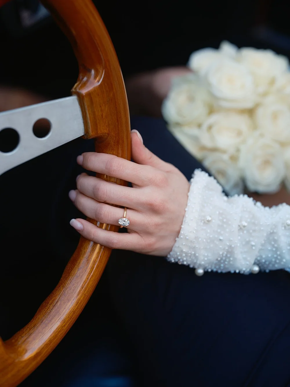 Detail of the bride's hand with her ring on the wooden steering wheel, with a white rose bouquet
