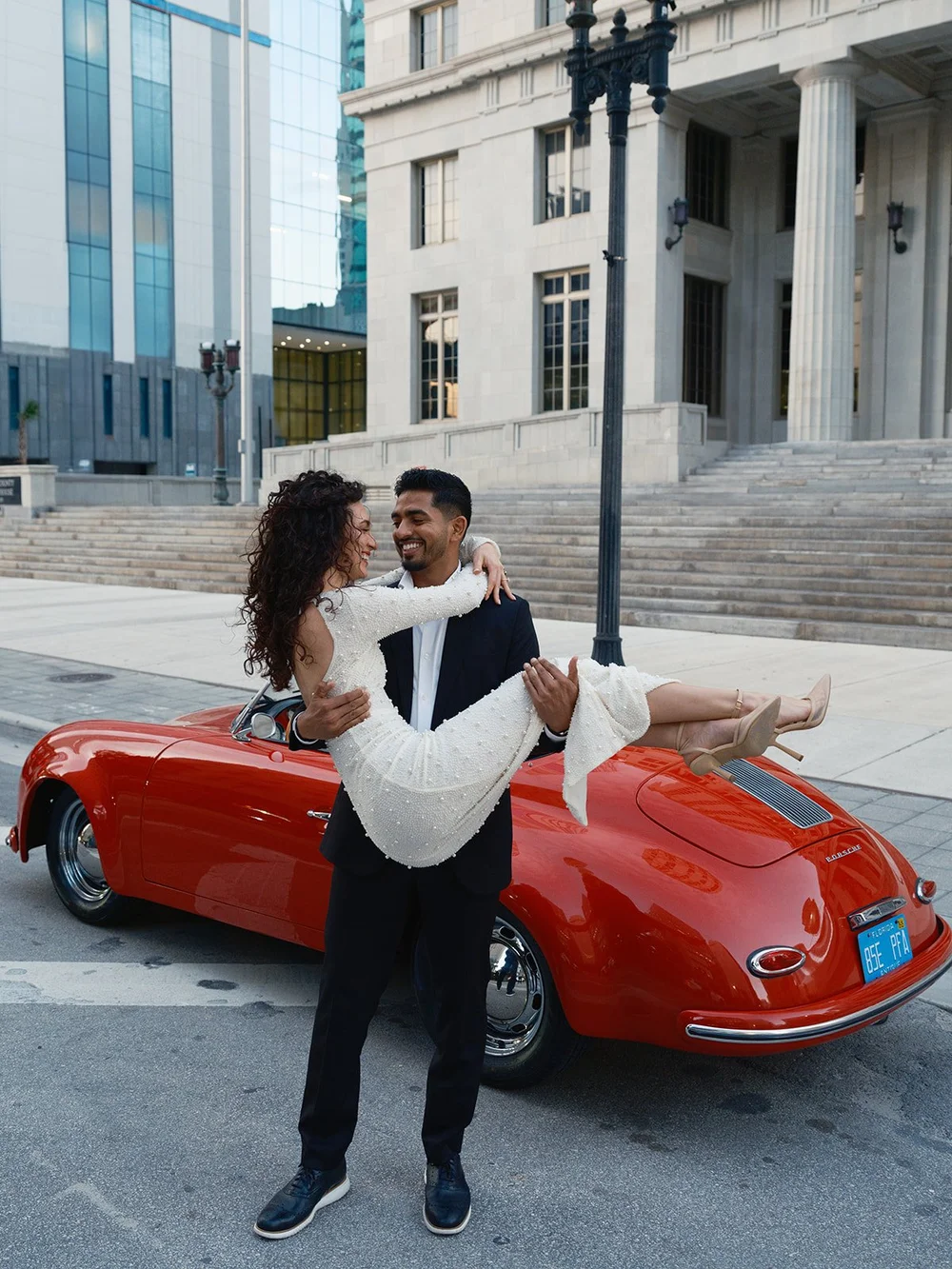 Groom lifting the bride in his arms in front of a red vintage convertible on a downtown Miami street