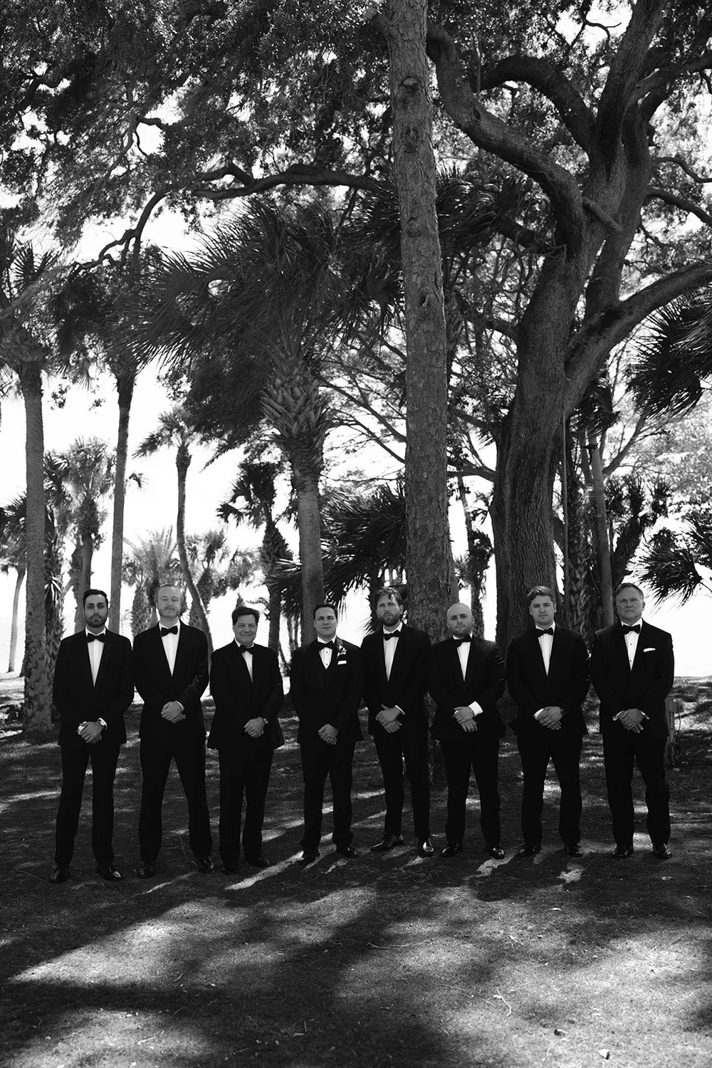 Black and white photo of the groomsmen standing in a line on the lawn under palm trees