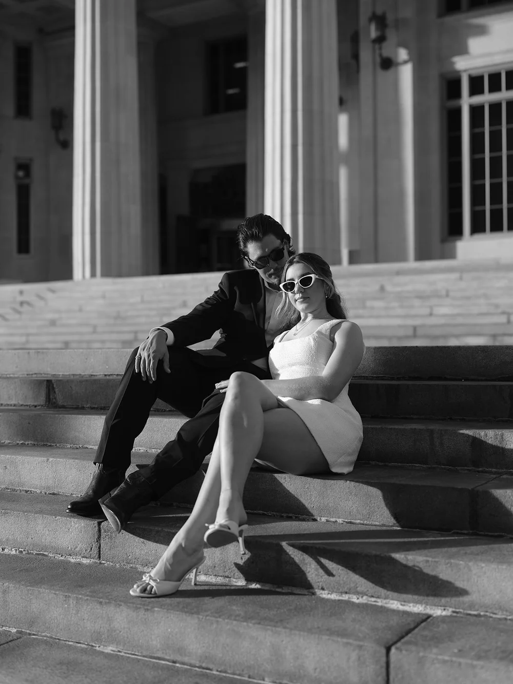 Black and white image of the bride in sunglasses sitting on the courthouse steps with the groom leaning back beside her