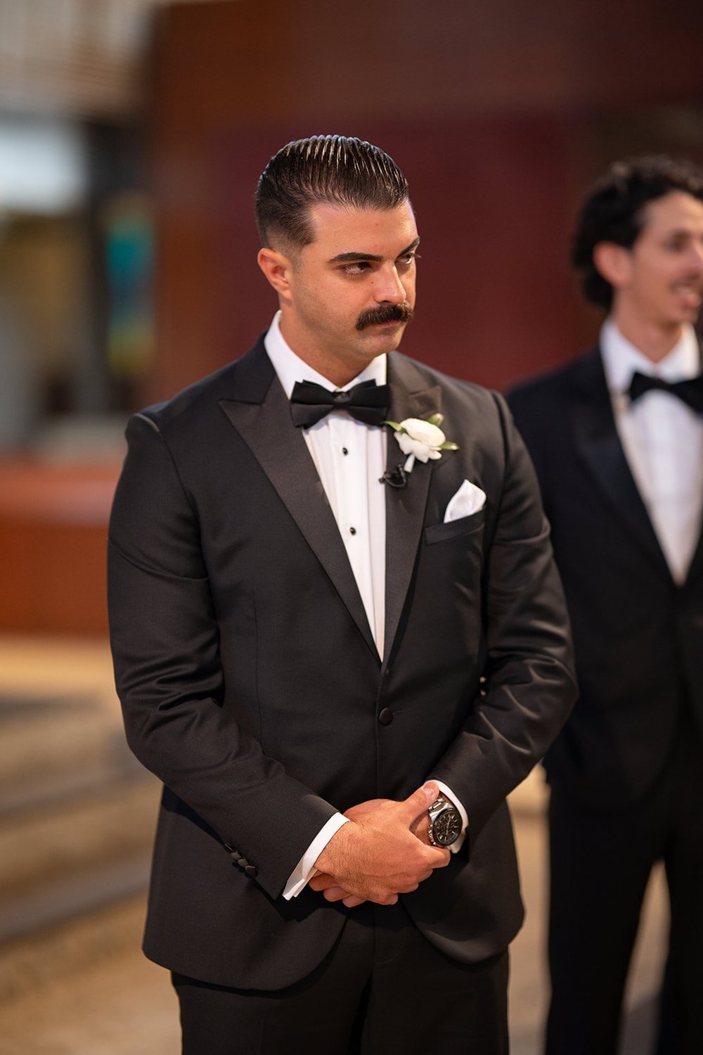 Austin in a black tuxedo with a serious expression as he waits at the altar with a groomsman behind him