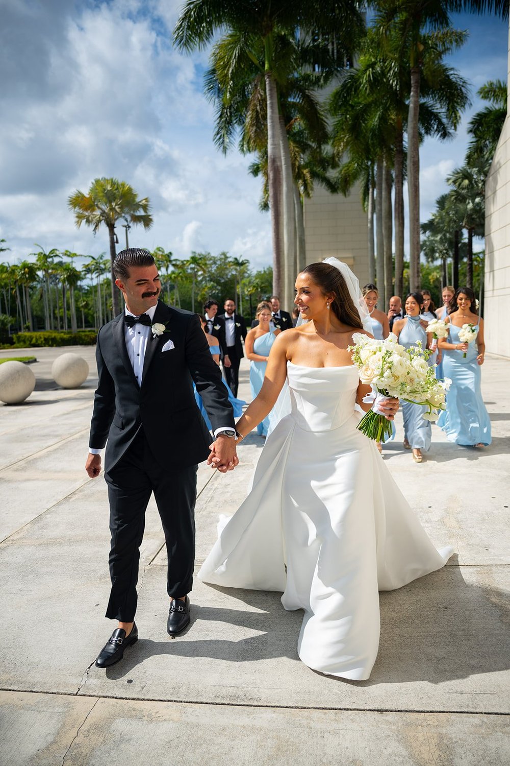 Rachel and Austin walking hand in hand on a path lined with palm trees, bridesmaids in blue behind them