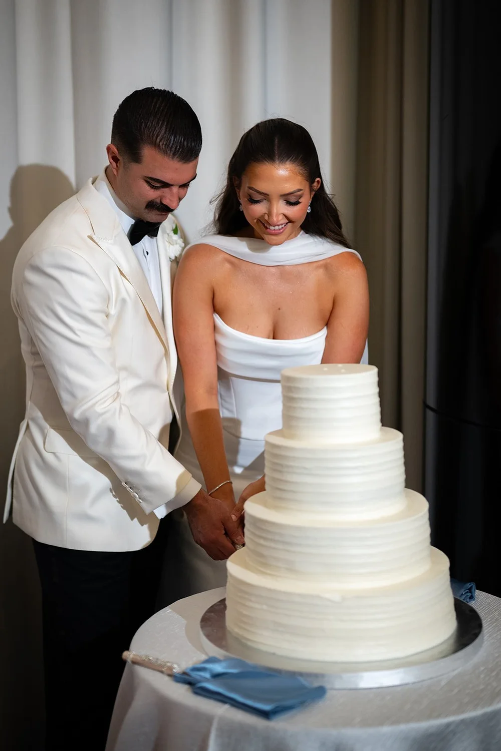 Austin in a white dinner jacket and Rachel cutting their tiered white wedding cake