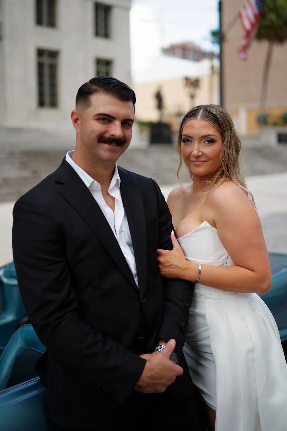 Portrait of Austin in a black suit and Rachel in a white dress next to the blue Mustang in downtown Miami