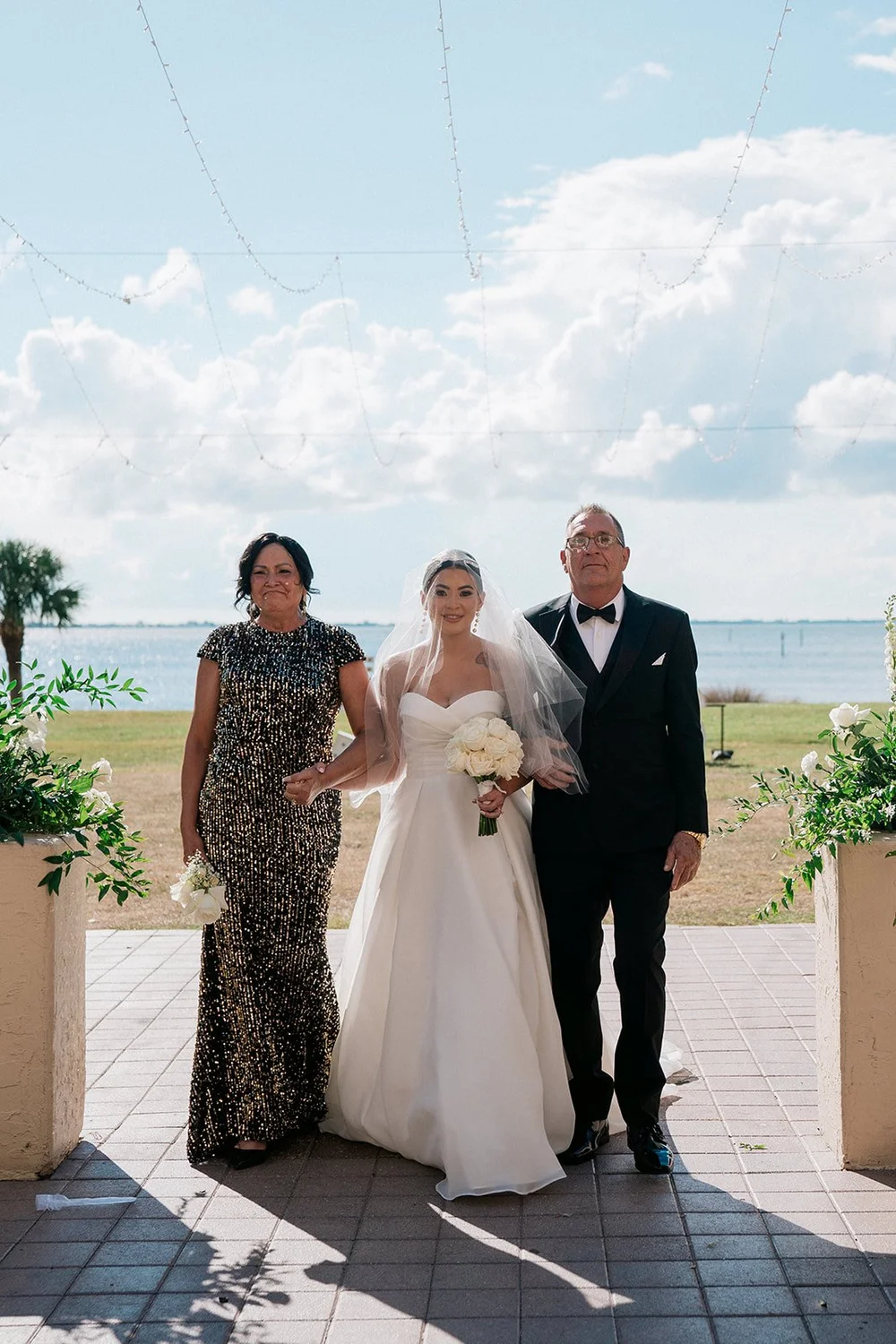 The bride between a woman in a gold dress and a man in a tuxedo on the estate terrace, with Sarasota Bay and clouds behind them