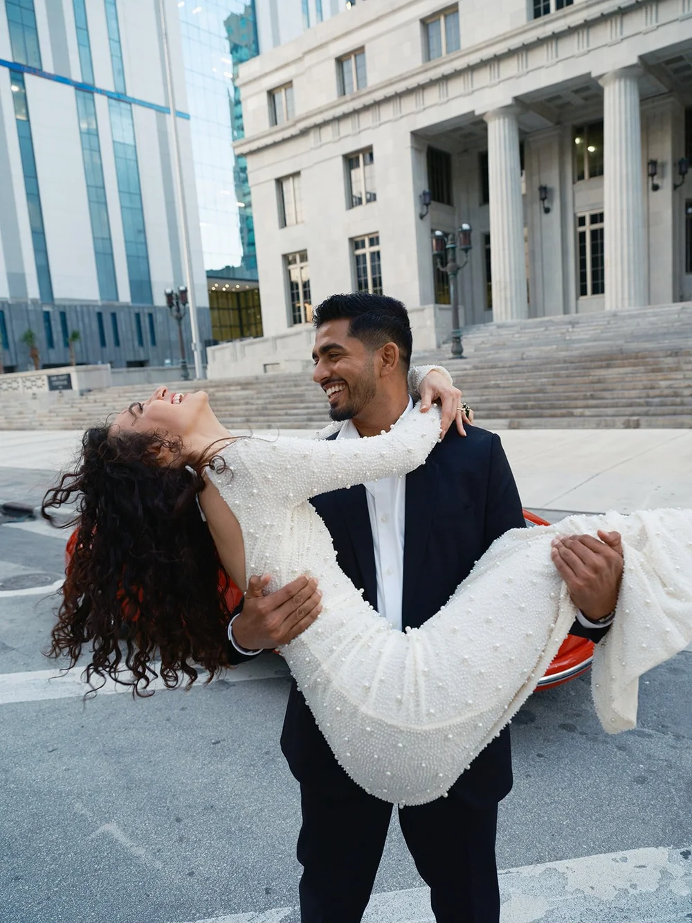 Groom carrying the bride as she leans back laughing, with the red car and courthouse behind them