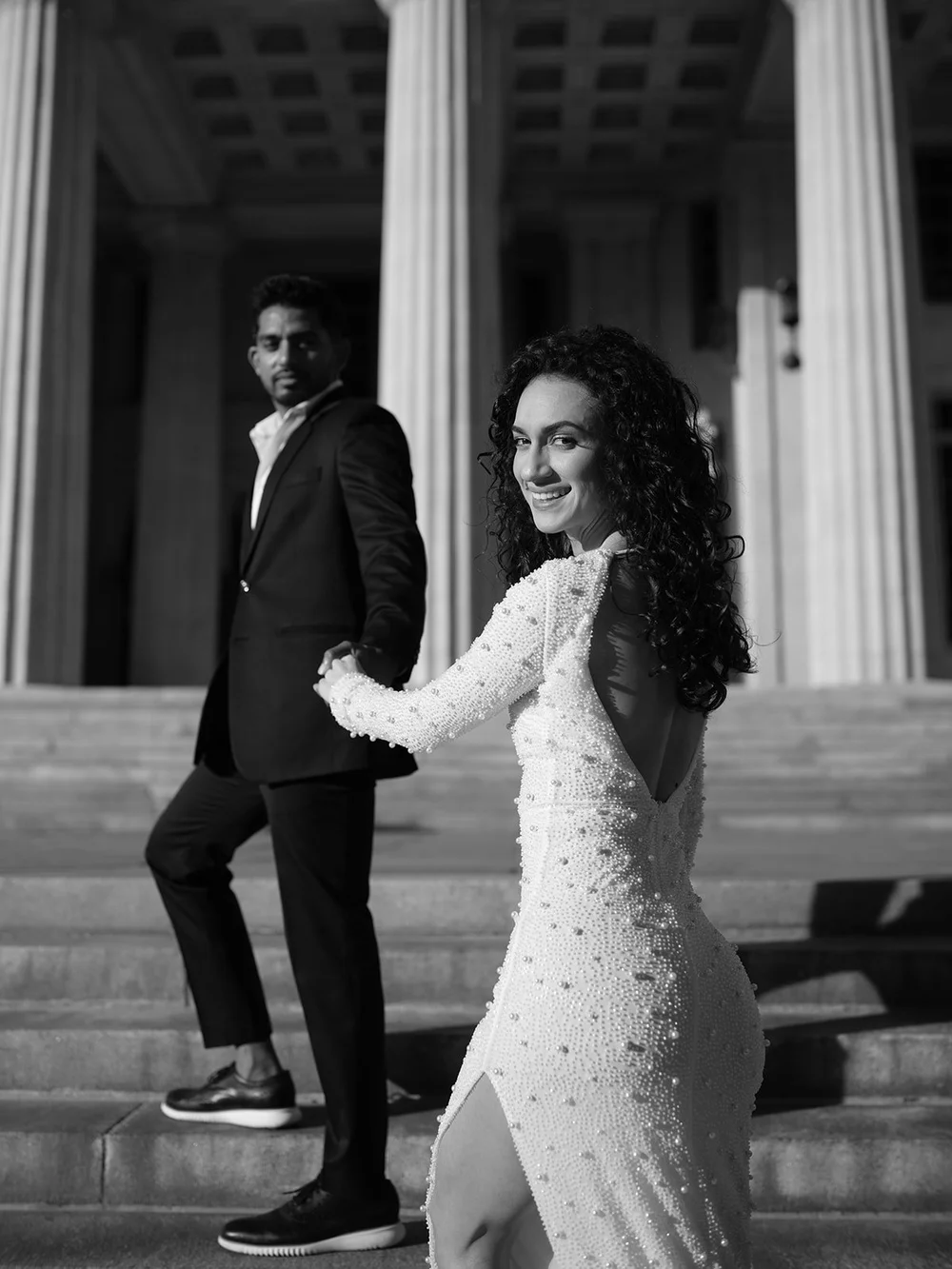 Black and white photo of the bride looking back at the camera as she walks up the courthouse steps with the groom behind her