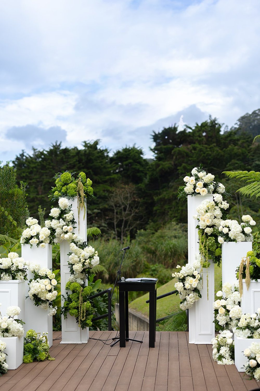 White flower arrangements on columns along the wooden ceremony deck with green mountains behind