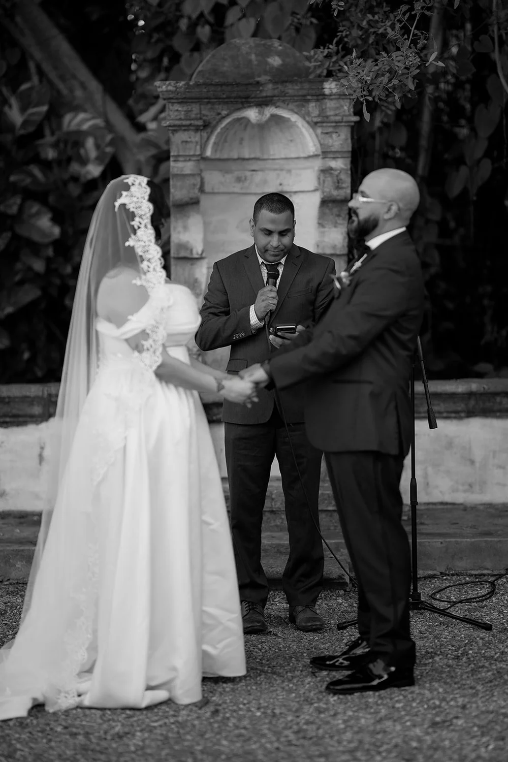 Maria and Christian facing each other in front of the officiant by a stone niche, in black and white