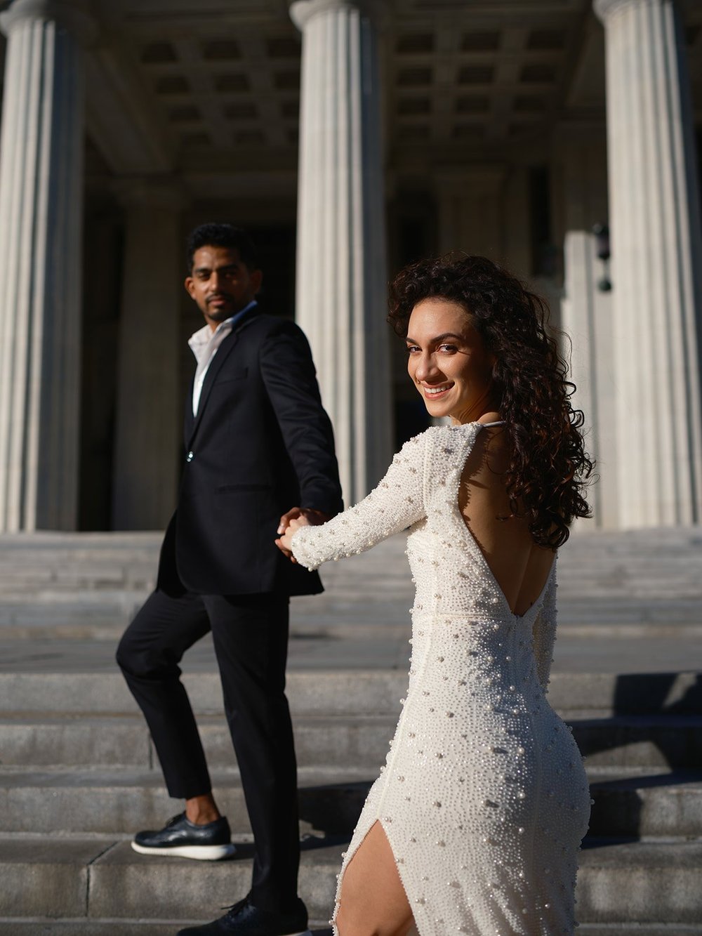 Bride in a backless white dress looking back at the camera with the groom beside her under tall courthouse columns