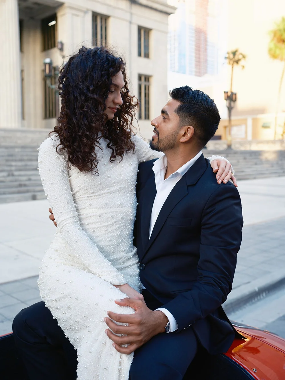 Bride sitting on the groom's lap on the red car, looking at each other