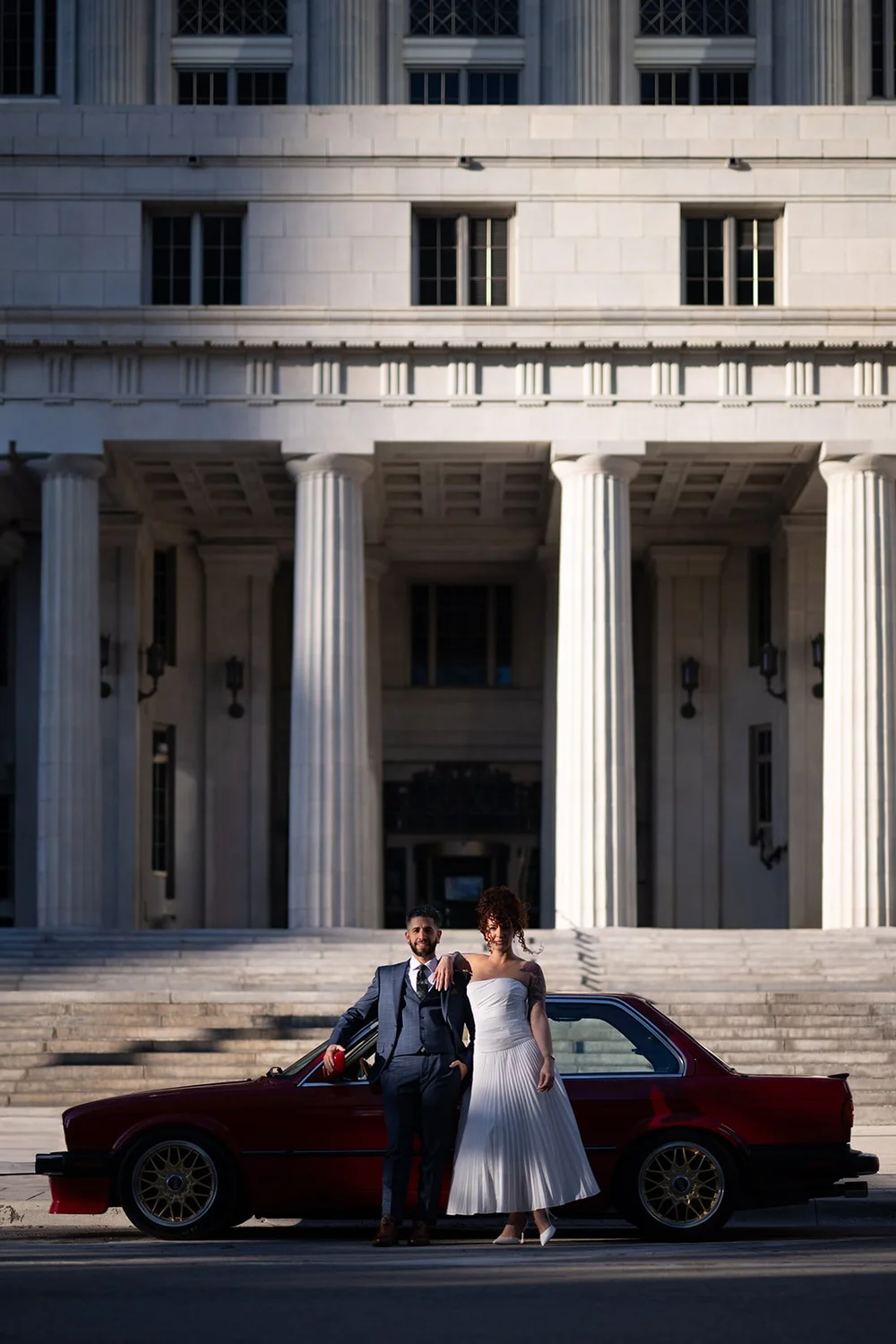 Bride and groom beside a red BMW in front of the columns of the Miami-Dade County Courthouse in downtown Miami