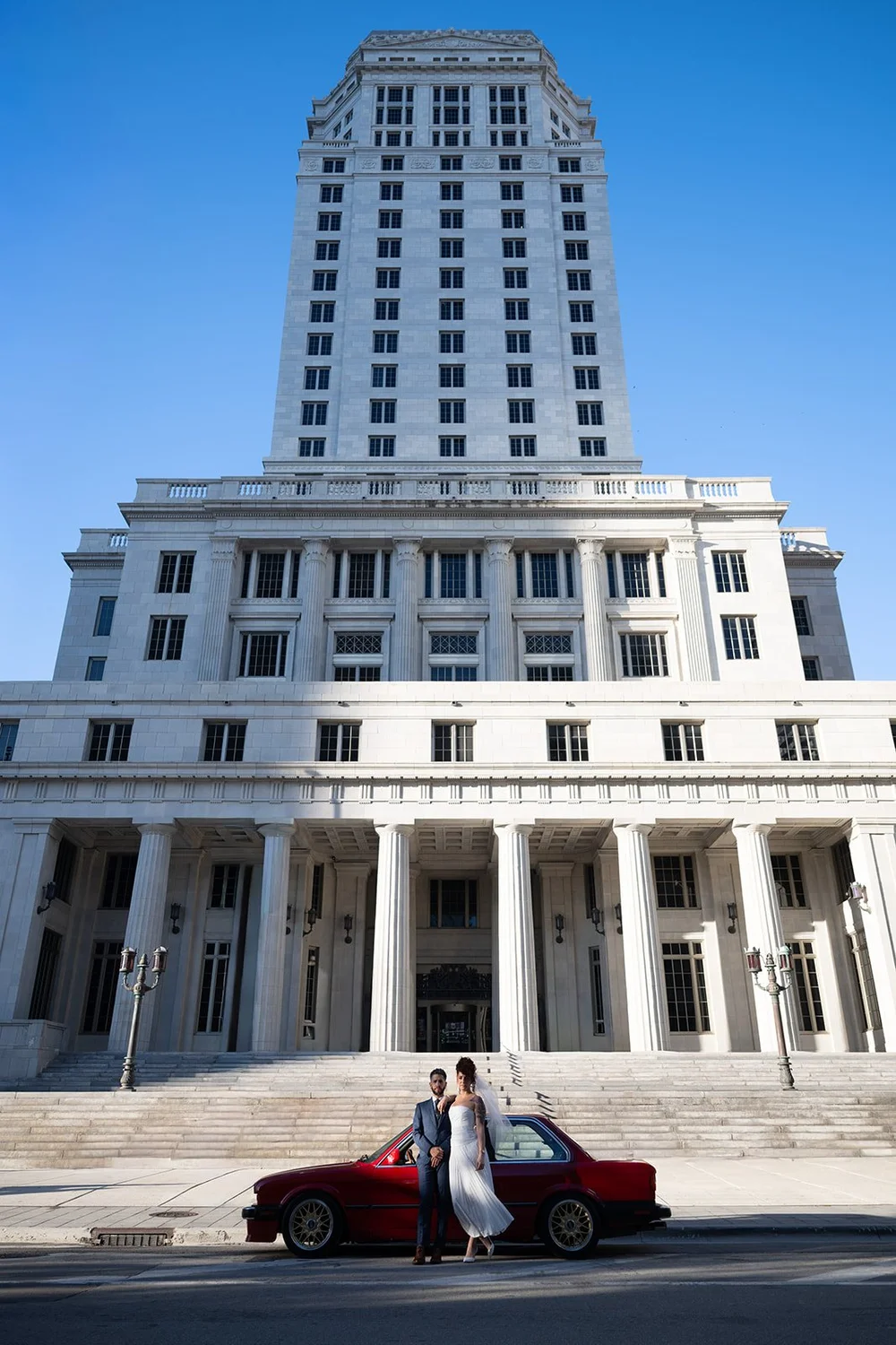 Bride and groom with a red BMW in front of the Miami-Dade County Courthouse tower in downtown Miami