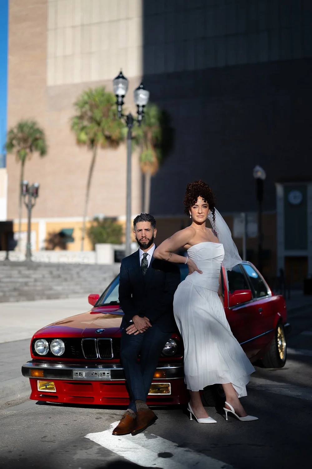 Groom leaning on the red BMW while the bride's veil blows in the wind