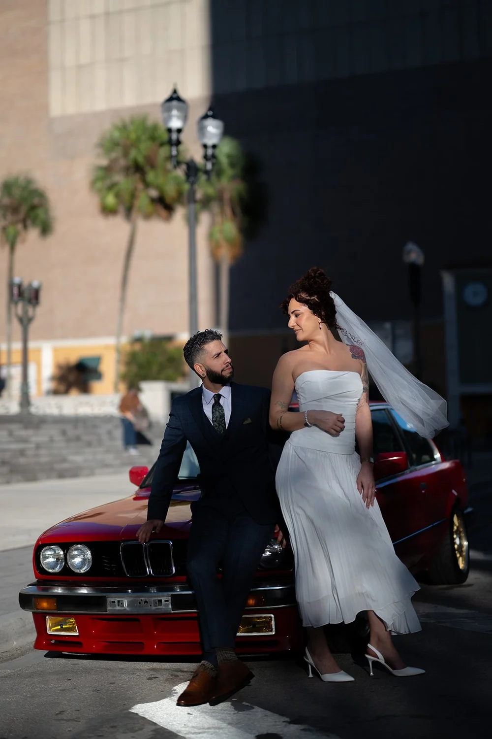 Bride in a white dress and veil beside the groom and a red BMW on a downtown Miami street