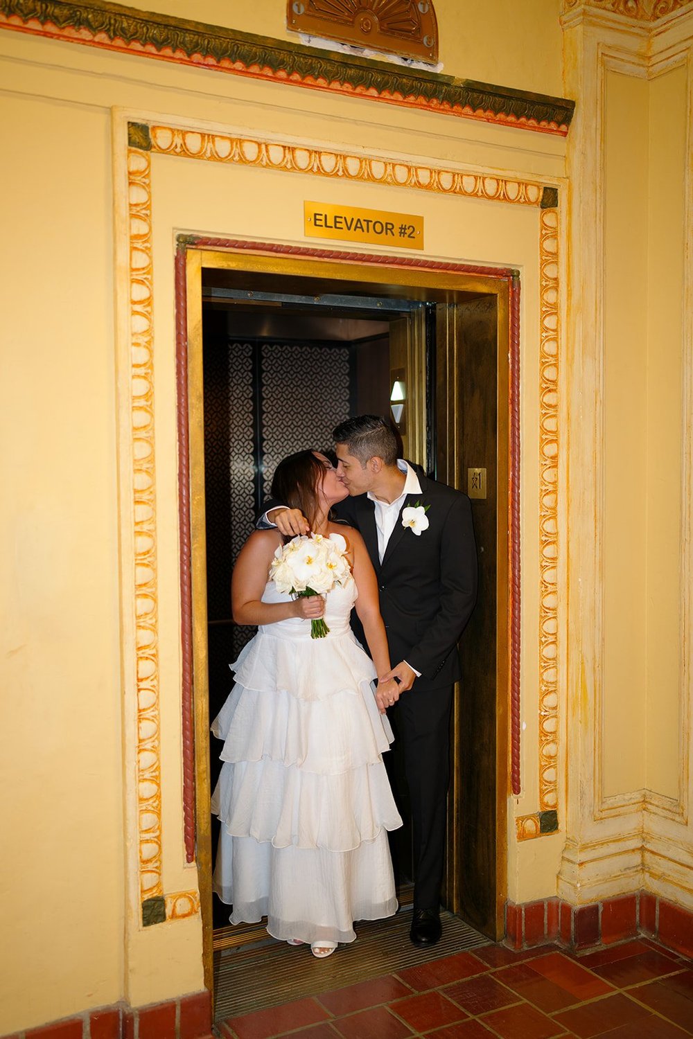 Chris and David standing in a gold-trimmed elevator doorway with warm yellow walls and a red tile floor