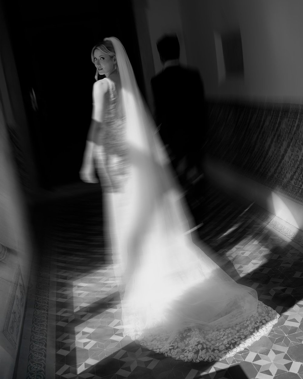 Black and white motion-blur photo of Ellen in her veil looking back over her shoulder as Jacob walks ahead of her down a tiled hallway
