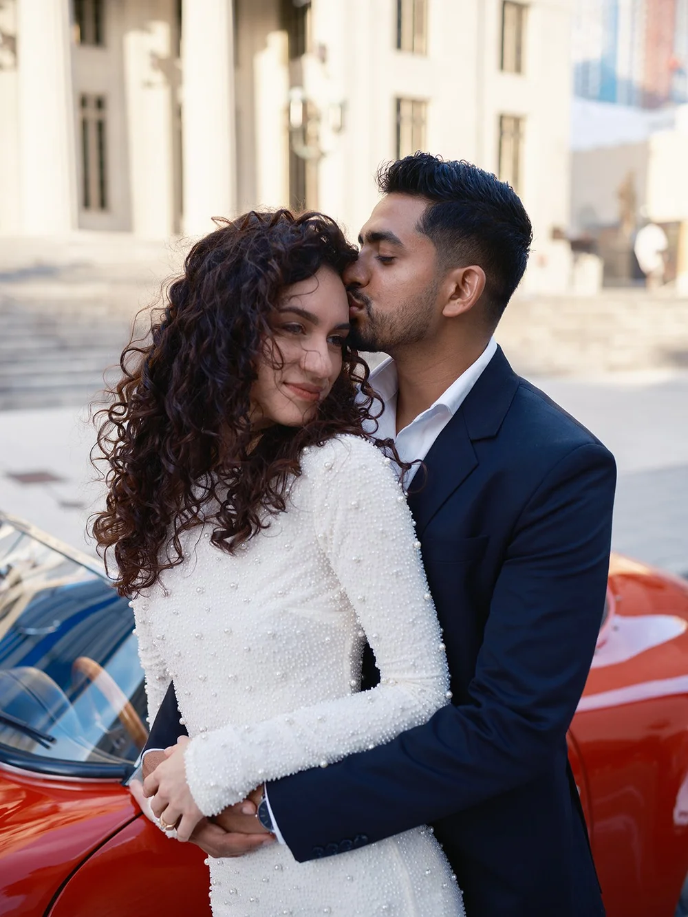 Groom kissing the bride's temple as she smiles, with the red convertible behind them