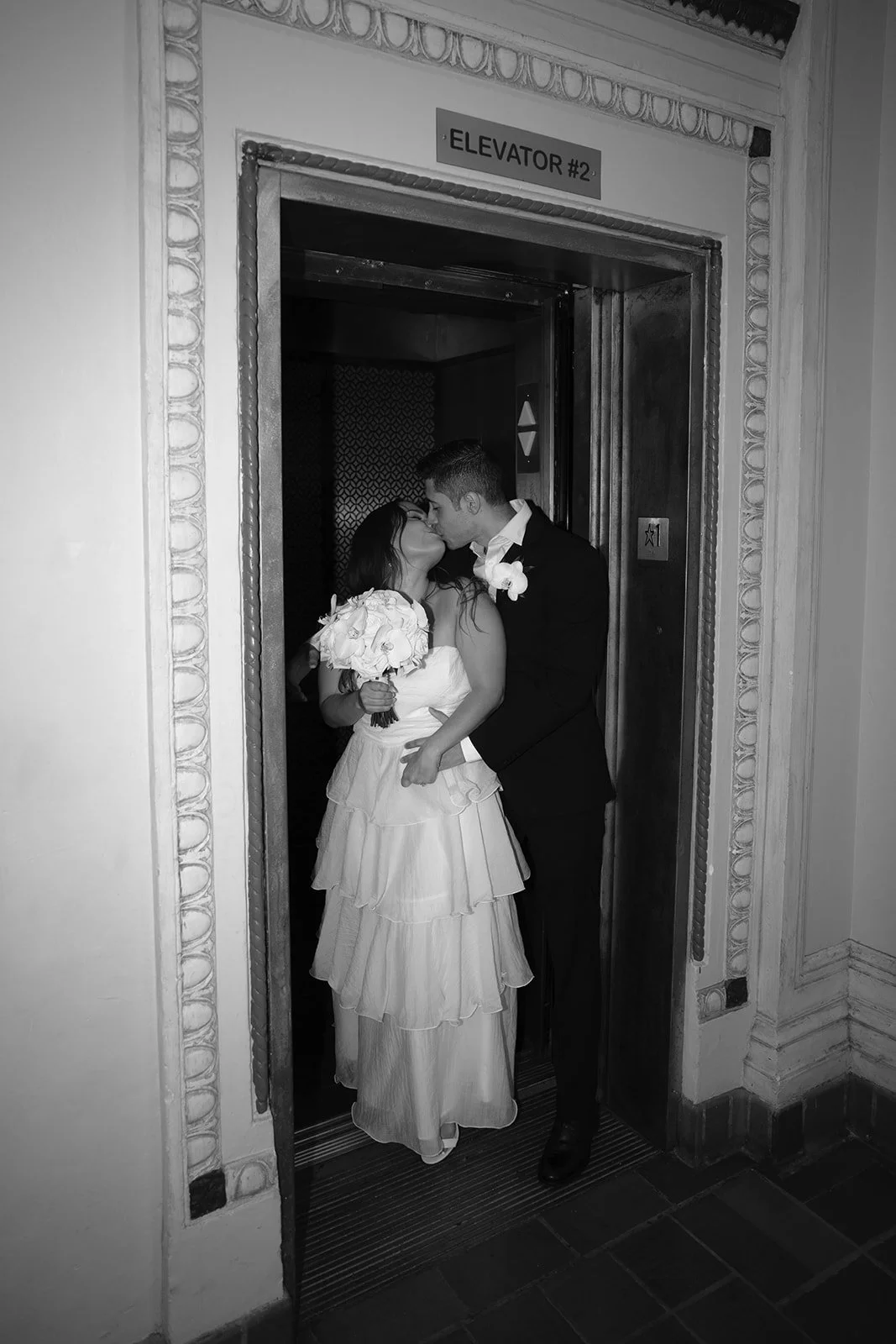Black and white flash photo of Chris and David kissing in an elevator doorway, Chris holding a white bouquet, for their black and white courthouse wedding photos