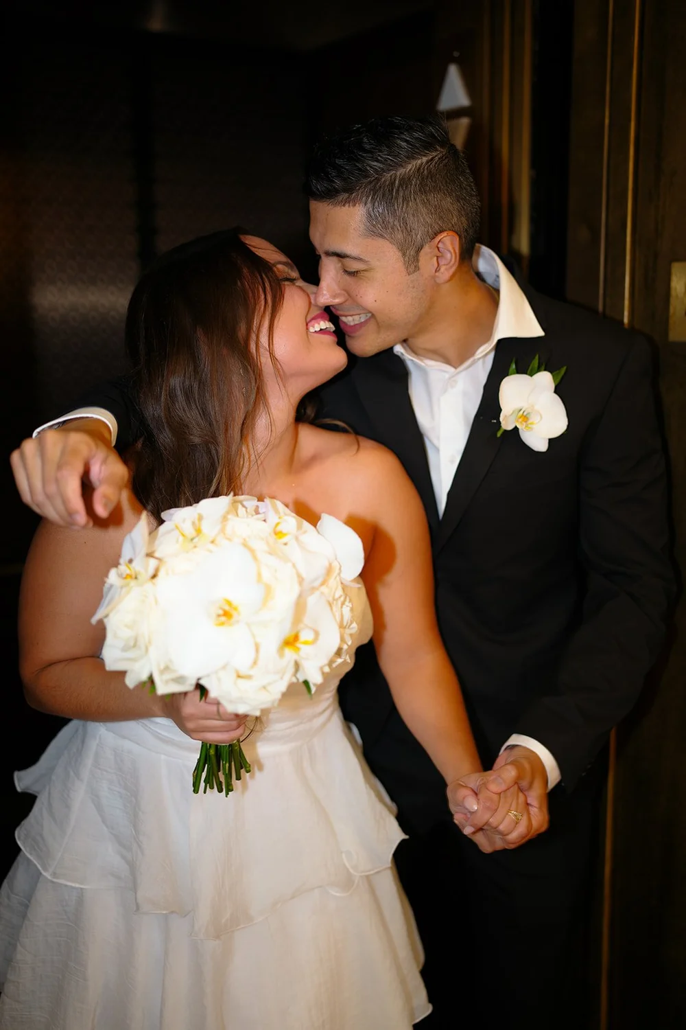 Color flash photo of David kissing Chris's cheek while she holds a white bouquet against a dark background