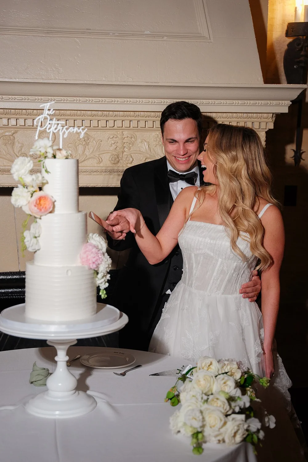Jane and Berkeley smiling beside a three-tier white wedding cake in front of a carved stone fireplace