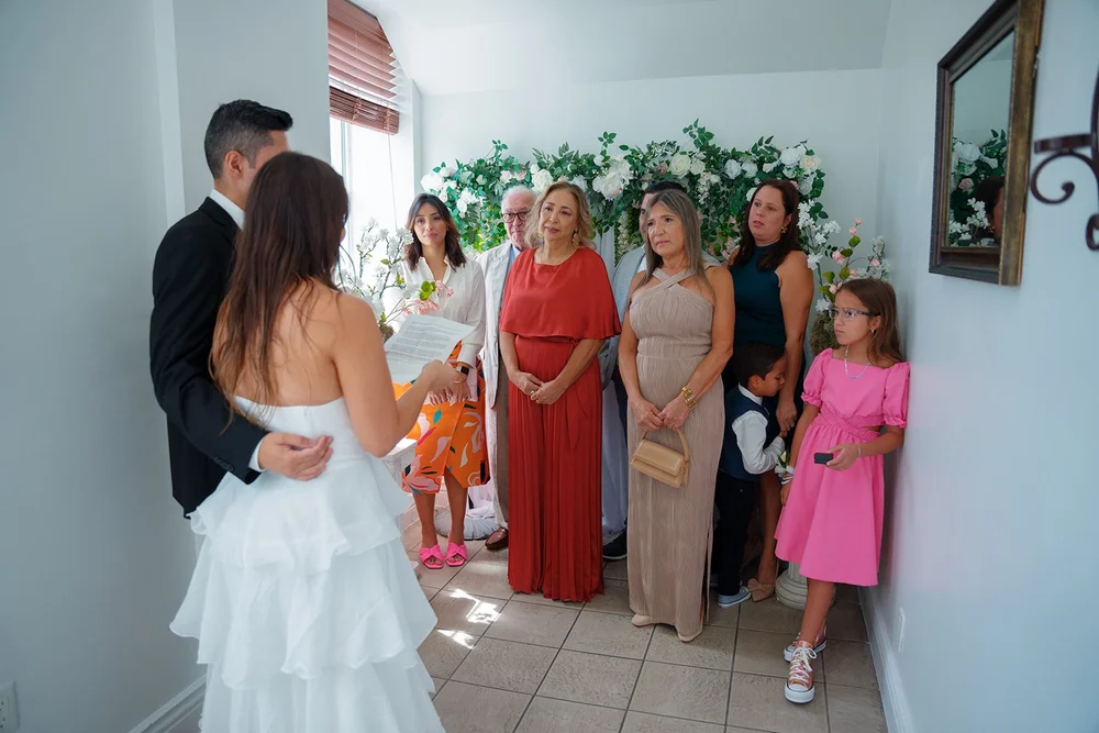 Wide frame of Chris and David standing together in front of their guests, with white flowers on the wall behind them