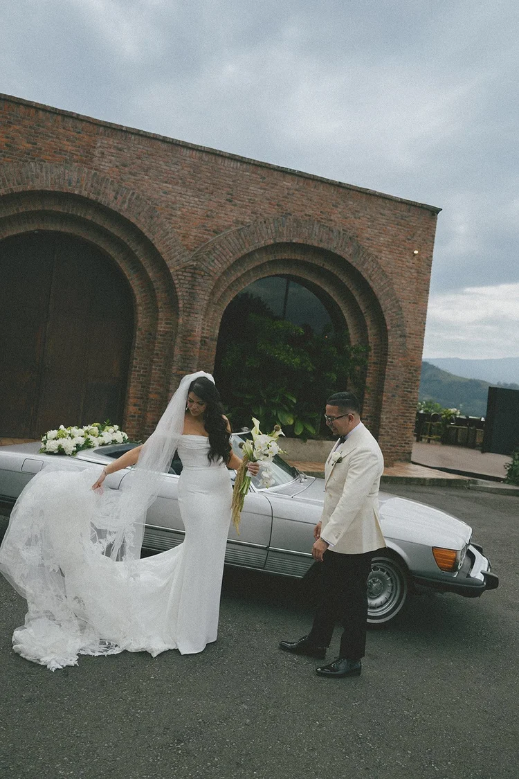 Film photo of Krystine in her veil and wedding gown beside a vintage Mercedes convertible in front of brick arches in Medellín as Douglas stands nearby