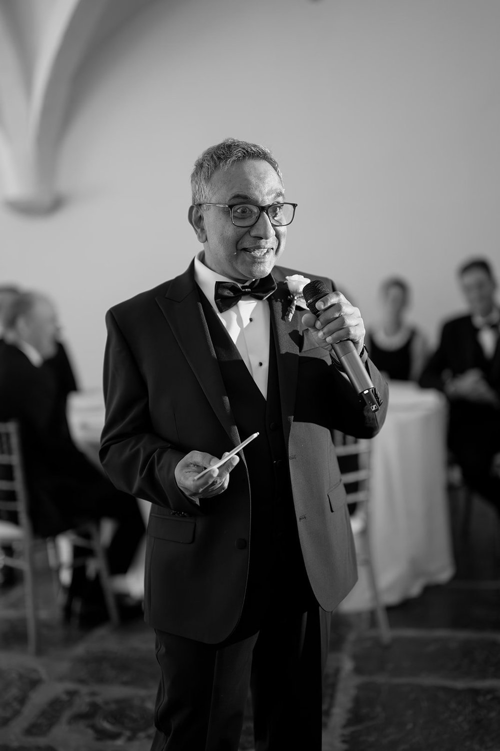 Black and white photo of a guest in a tuxedo giving a toast with a microphone