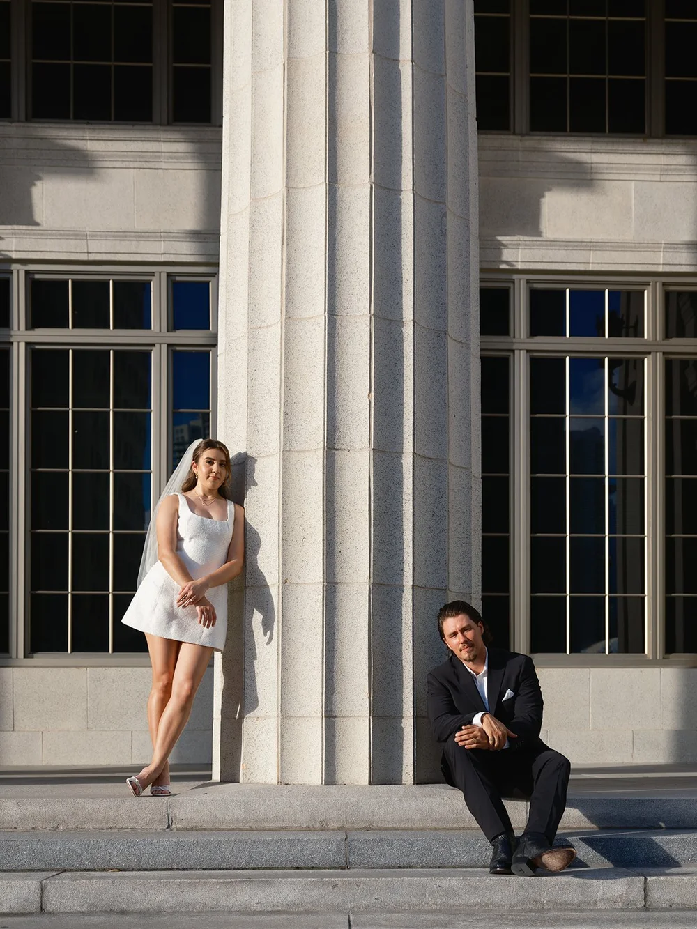 Bride leaning on a stone column in a short white dress while the groom sits on the steps