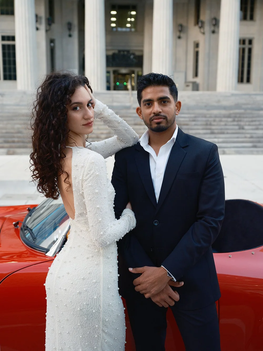 Bride in a white beaded dress and groom in a navy suit beside a red vintage convertible with the courthouse steps behind them