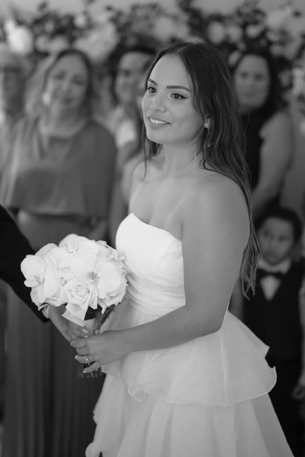 Black and white image of Chris holding her white bouquet with guests standing behind her