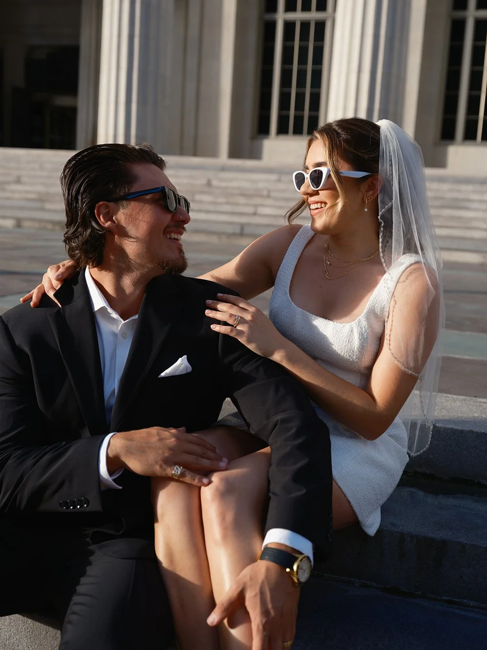 Groom and bride in sunglasses sitting together on the courthouse steps, smiling at each other