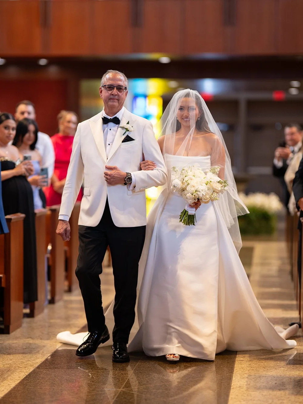 Rachel in her white gown and veil walking down the church aisle with a man in a white dinner jacket while guests look on