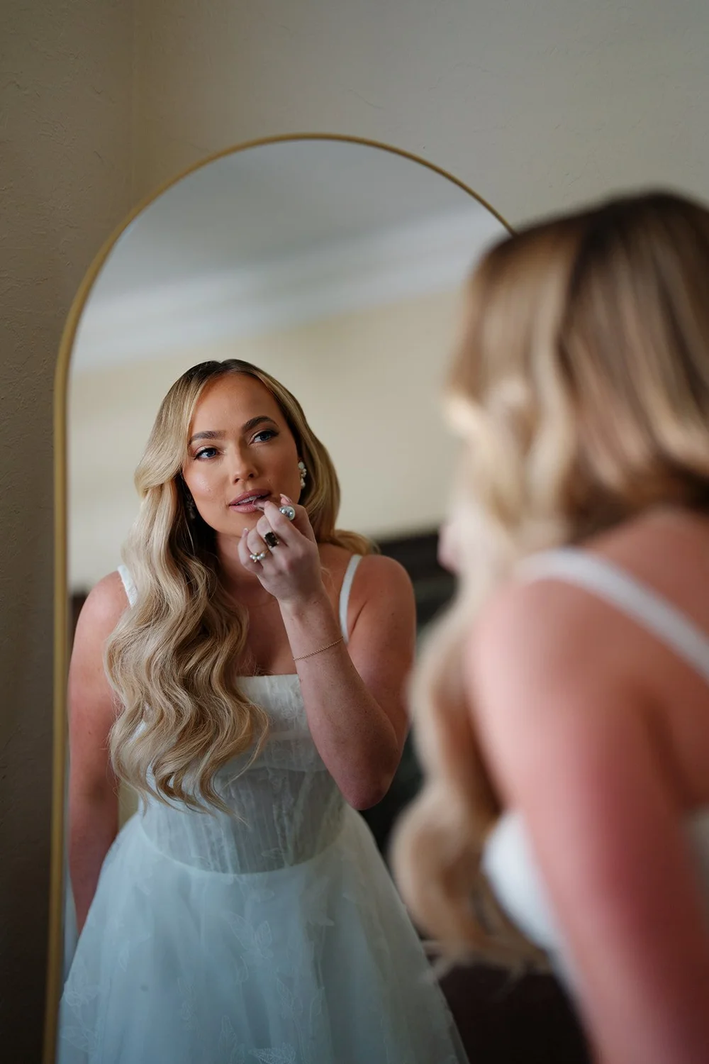 Jane in her wedding dress looking in a mirror with her hand near her face while another woman stands beside her